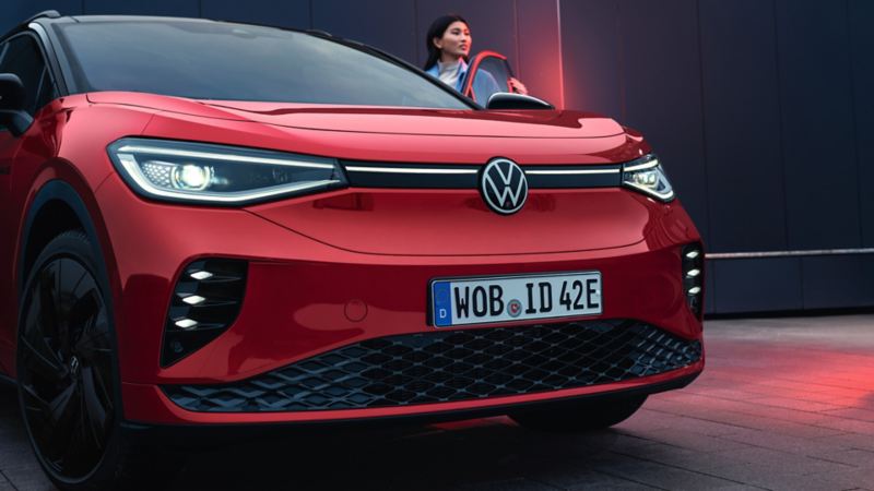 Ένα VW ID.4 GTX σε κόκκινο χρώμα με ανοιχτή πόρτα οδηγού. Μία γυναίκα στέκεται στην πόρτα και κοιτάζει προς τα εμπρός.