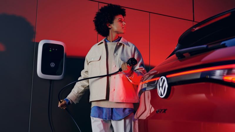 Une VW ID.4 GTX rouge vue de l’arrière. Une femme tient le câble de recharge de l’ID.Charger dans ses mains et sourit en direction du véhicule.