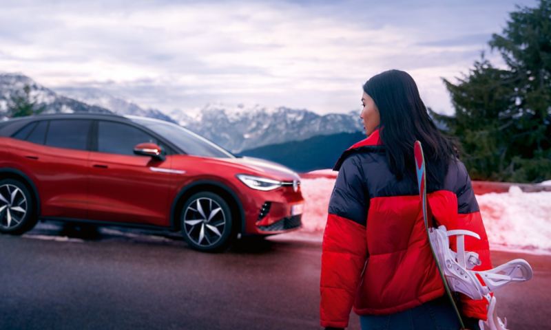 VW ID.4 GTX visible de côté en rouge. Sur le bord de la route en face des montagneux, une femme avec un snowboard se dirigeant vers elle