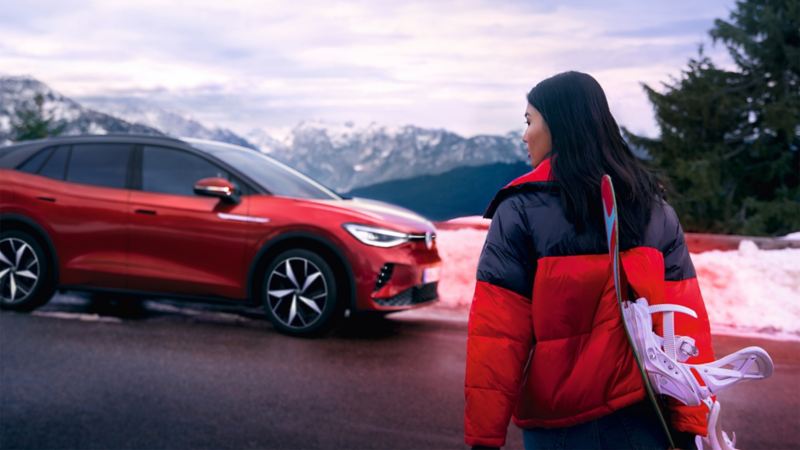 Zijaanzicht van een rode VW ID.4 GTX tegen een bergachtergrond, op de voorgrond kijkt een vrouw in de richting van de auto
