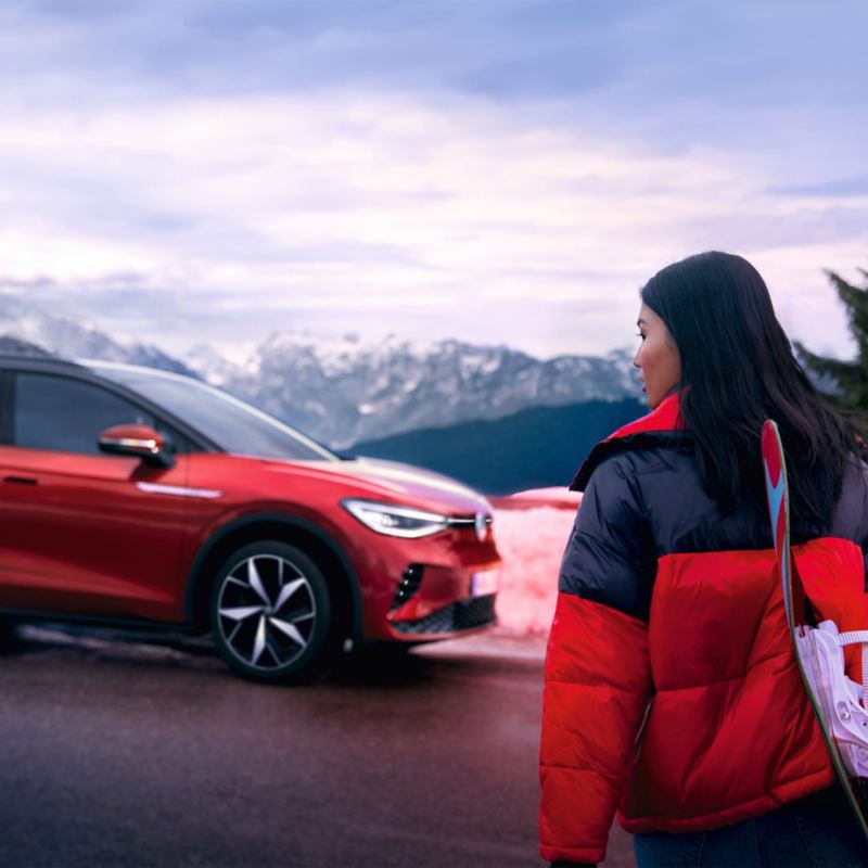 Zijaanzicht van een rode VW ID.4 GTX tegen een bergachtergrond, op de voorgrond kijkt een vrouw in de richting van de auto.