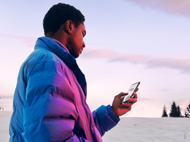 Ein Mann in Winterjacke steht in verschneiter Landschaft und schaut auf sein Smartphone.