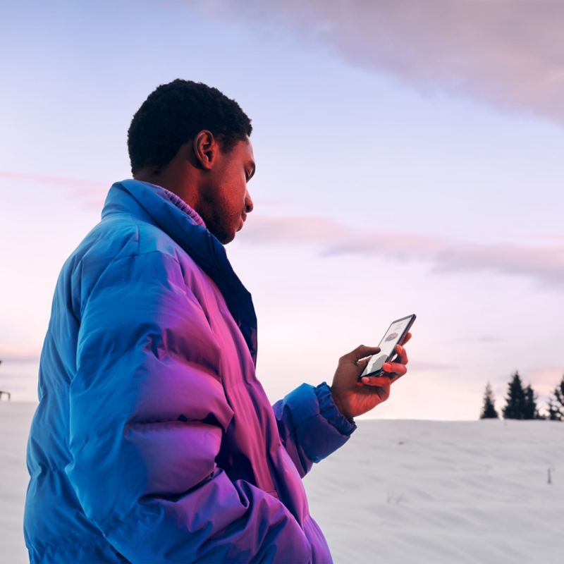 Een man met een winterjas aan staand in een besneeuwd landschap en kijkend op zijn smartphone.