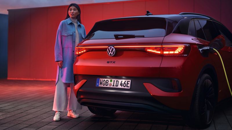 Arrière d'un VW ID.4 GTX stationné en charge avec les feux arrière allumés et une femme debout à côté