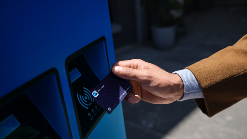 Eine Hand hält eine VW We Charge Ladekarte an eine blaue Ladestation.