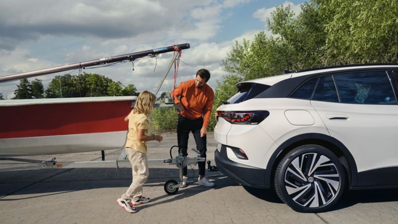Vue latérale de la Volkswagen ID.4 blanche. Un homme et une jeune fille attachent un bateau à une remorque sur le dispositif d’attelage.