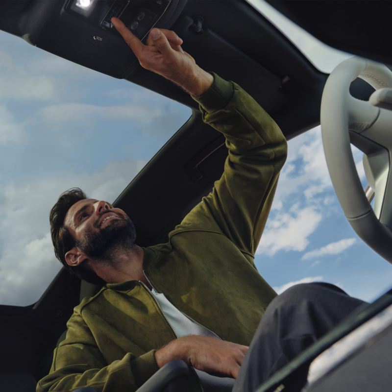 Vaizdas iš apačios į „VW ID.4“ elektromobilio su panoraminiu stikliniu stogu prietaisų skydelį, vairuotojo sėdynėje sėdintis vyras naudojasi viršutinėje konsolėje esančiais valdikliais