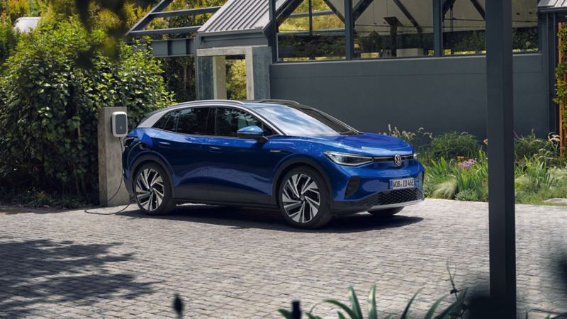 Ein blauer VW ID.4 parkt seitlich vor einem grauen, modernen Haus und lädt an der ID. Charger Wallbox. Im Hintergrund viel grün.