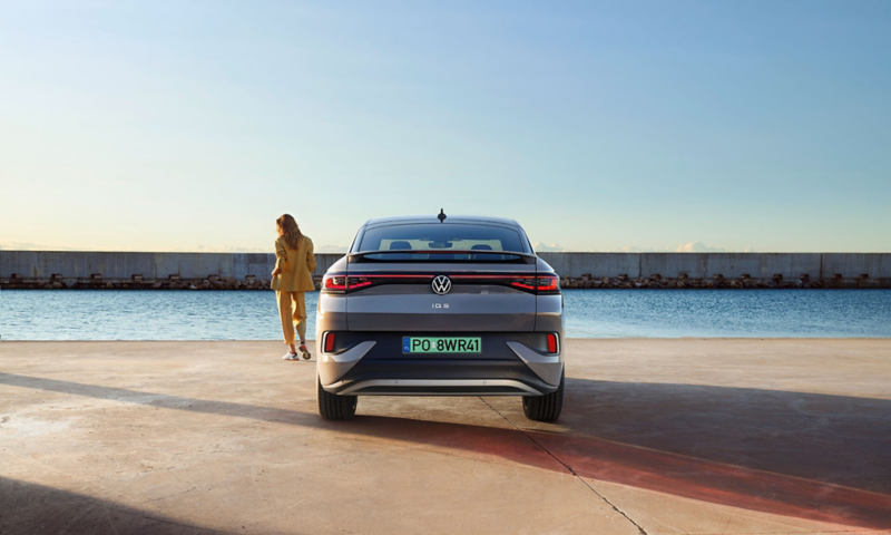 Samochód elektryczny Volkswagen ID.4 zaparkowany nad wodą, z osobą w żółtej odzieży obok podczas zachodu słońca