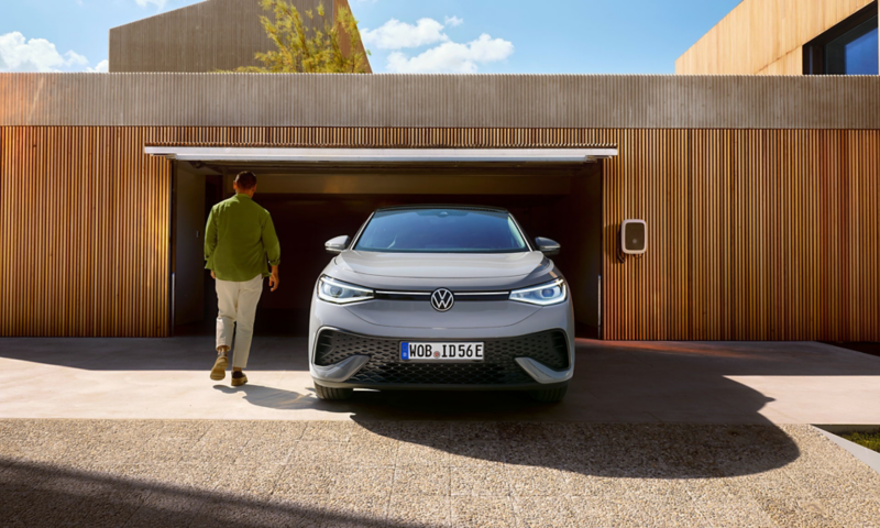 Front view of a silver VW ID.5 in front of an open garage with a wallbox, a man is walking towards the garage door. 