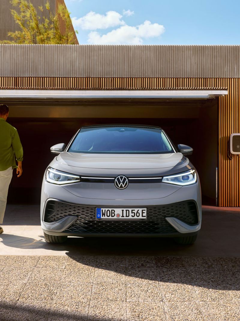 Frontansicht eines silberfarbenen VW ID.5 vor einer geöffneten Garage mit Wallbox, ein Mann geht auf das Garagentor zu.