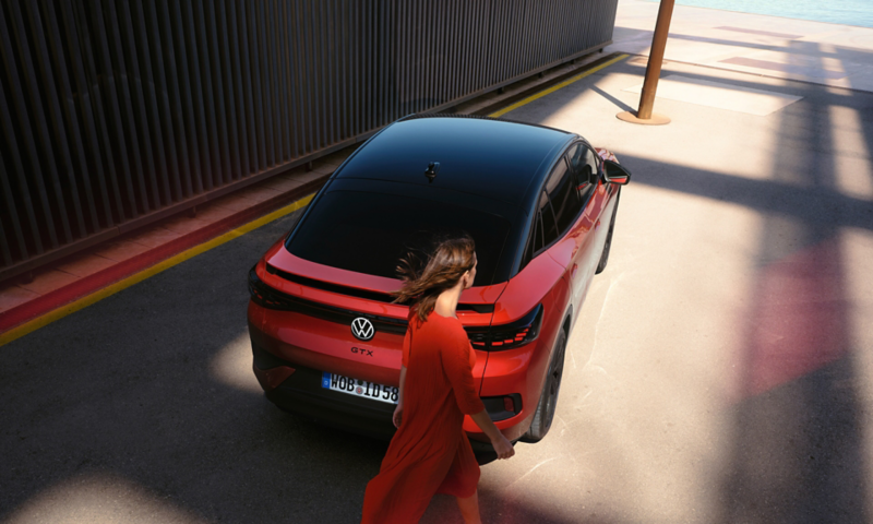Ένα κόκκινο VW ID.5 GTX σε μία πάροδο. Άποψη από επάνω στην οροφή και στο πίσω μέρος. Μία νέα γυναίκα περνάει από δίπλα.