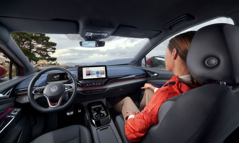 Intérieur d'un Volkswagen ID.5 GTX avec le système infotainment allumé sur Apple Music et une jeune femme installée place passager qui observe les montagnes.