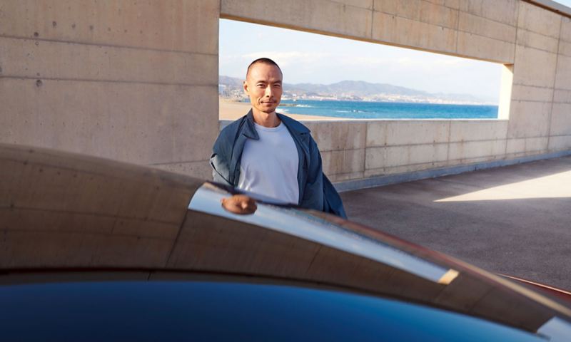 Мужчина стоит перед бетонной стеной, на заднем плане окно с видом на побережье. На переднем плане крыша VW ID.5 GTX