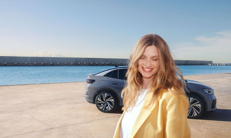 Young woman stands smiling in front of a gray VW ID.5 parked at a port.