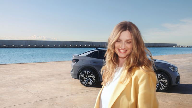 Vista lateral de un VW ID.5 gris en un muelle del puerto, con una mujer sonriente y vestida de forma brillante en primer plano