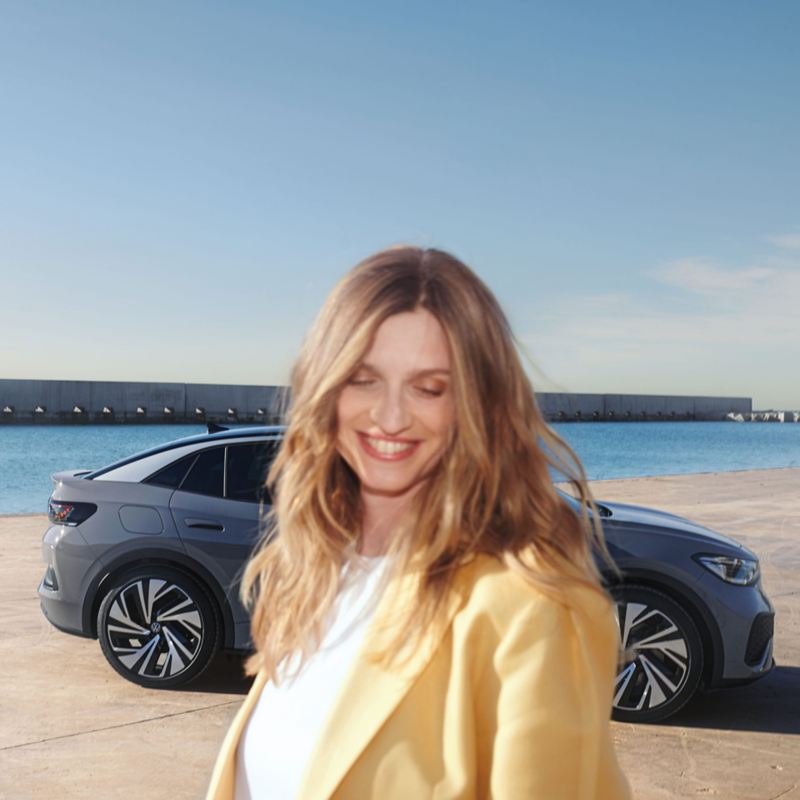Vista lateral de un VW ID.5 gris en un muelle del puerto, con una mujer sonriente y vestida de forma brillante en primer plano.