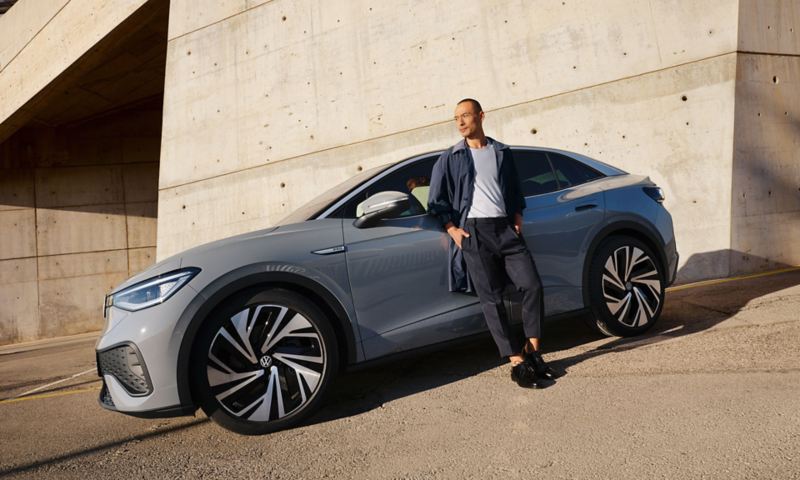 Side view of a gray VW ID.5 in front of a wall, a man leans against the driver's door. 