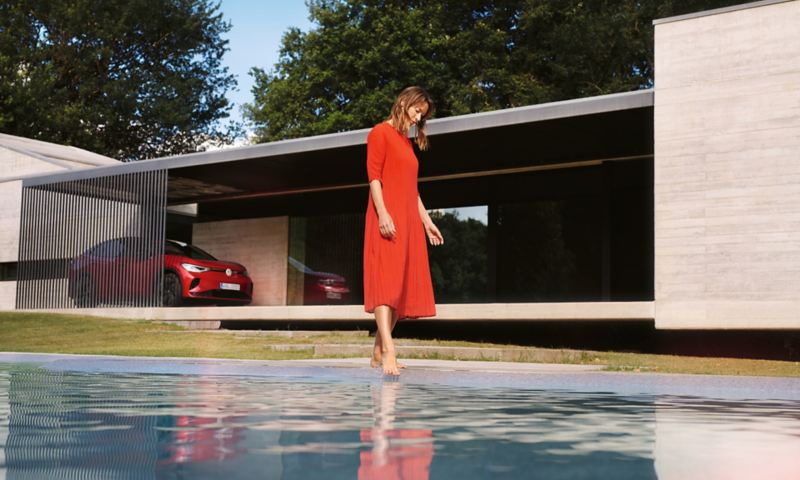 Une VW ID.5 GTX rouge est garée devant une maison. Une femme en robe rouge est au bord de la piscine.