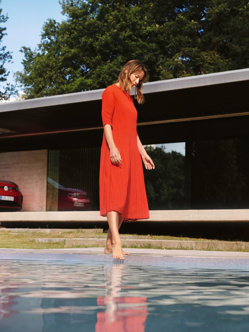 Raudonas Volkswagen ID.5 GTX stovi prie namo. Moteris, vilkinti raudona suknele, stoviniuoja prie baseino.