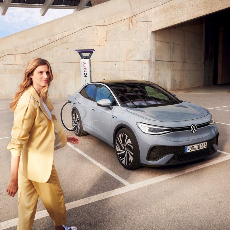 En grå VW ID.5 står på en parkeringsplats och laddas vid en laddstolpe, fronten och sidan är synlig, en kvinna går mot bilen