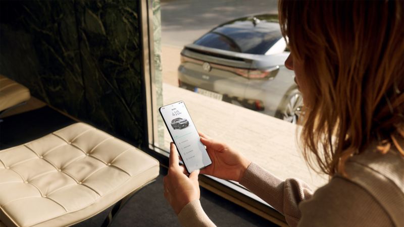Pilkos spalvos „VW ID.5“ vaizdas iš galo, pirmame plane moteris naudojasi išmaniuoju telefonu