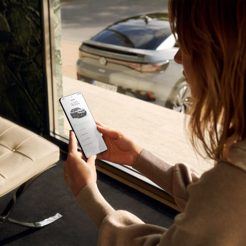 Achteraanzicht van een grijze VW ID.5, op de voorgrond bedient een vrouw een smartphone