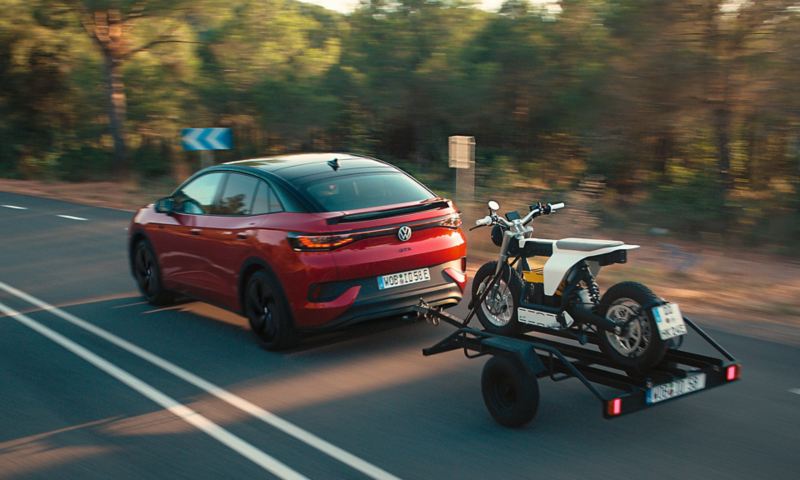 Une VW ID.5 GTX rouge roule de manière dynamique sur une route et tire une remorque avec une moto cross.