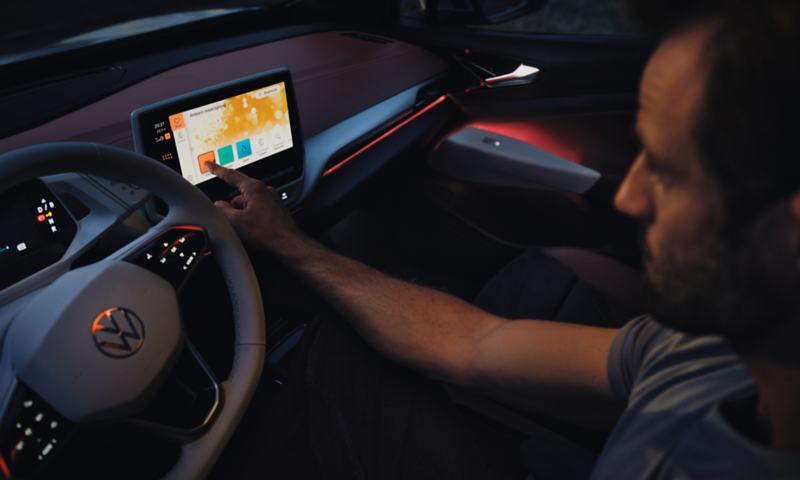 Vue intérieure de la VW ID.4, l'éclairage d'ambiance de l'habitacle s'allume en rouge. L'homme au volant utilise l'écran tactile.