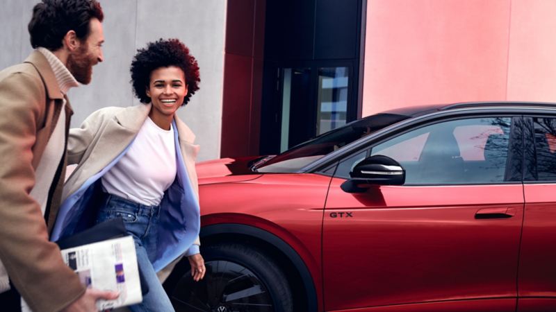 Rode VW ID.4 GTX geparkeerd aan de straatkant, van opzij gezien. Vrouw en man lopen lachend langs het voertuig.