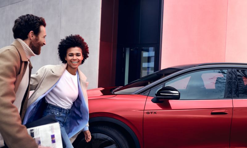 Une VW ID.4 GTX en rouge garée de côté en bordure de route. Une femme et un homme passent devant, le sourire aux lèvres.