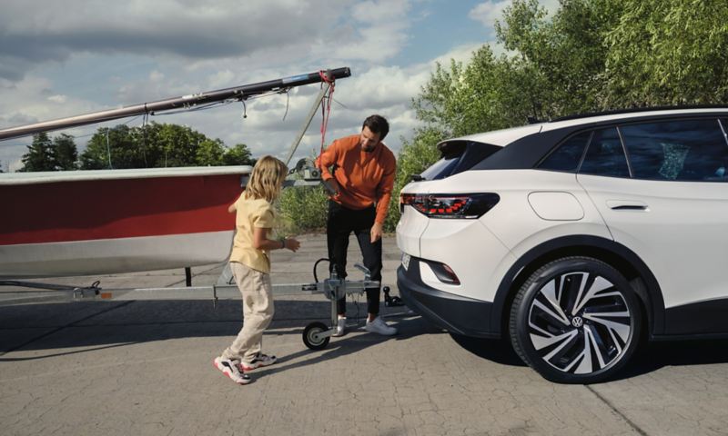 Vue latérale de la VW ID.4 blanche. Un homme et une jeune fille attachent un bateau sur une remorque au dispositif d'attelage.