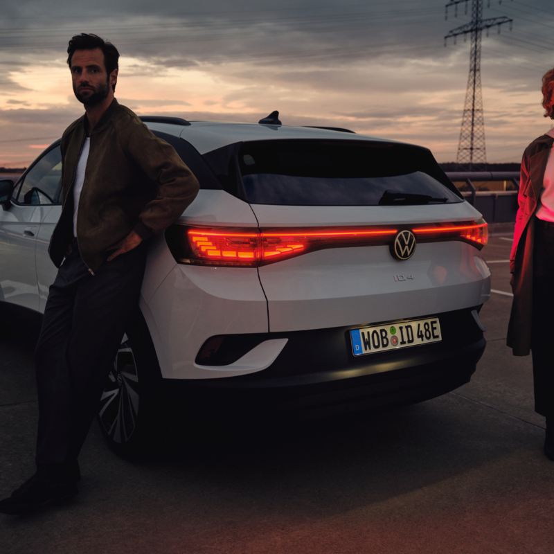 Un uomo e una donna al tramonto sostano ai lati di una Volkswagen ID.4 vista da dietro, per 3/4.