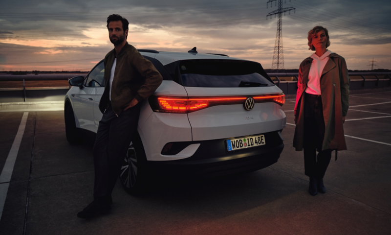 Un uomo e una donna al tramonto sostano ai lati di una Volkswagen ID.4 vista da dietro, per 3/4.