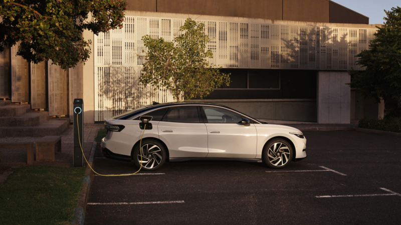 Ein weißer VW ID.7 in Seitenansicht lädt an einer Ladestation vor einem modernen Gebäude.