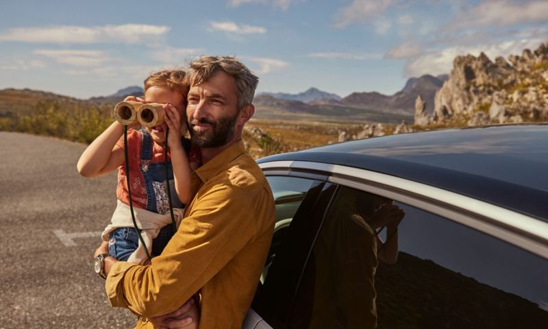A man is holding a child who is looking through binoculars. Both are in front of the VW ID.7.