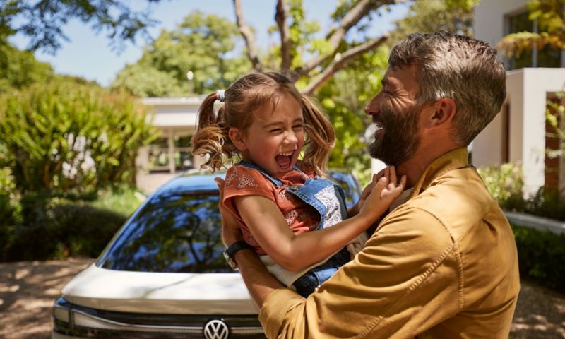 A man has a laughing child in his arms, the front of a VW ID.7 can be seen in the background.