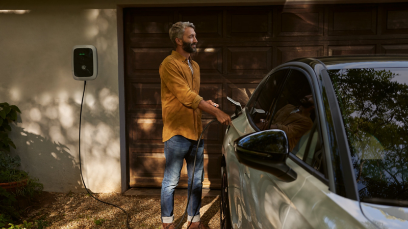 Ein Mann lädt einen weißen VW ID.7 Pro an einer Wallbox vor einem Haus.