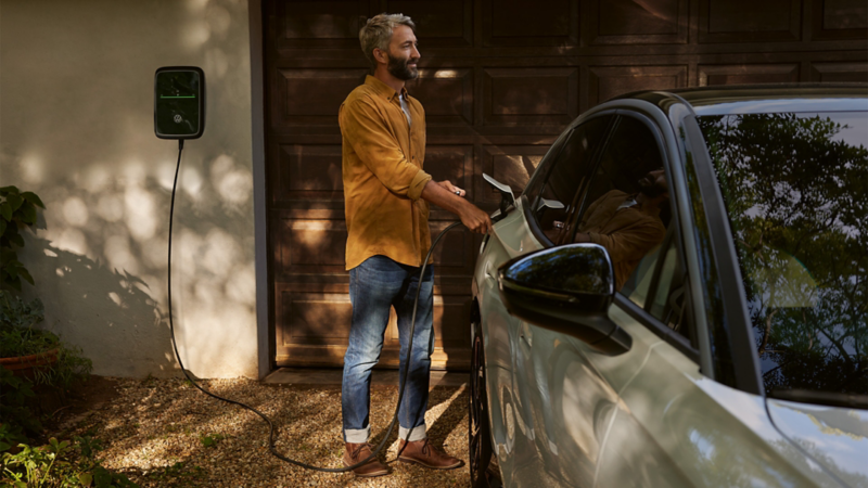 Ein Mann lädt einen weißen VW ID.7 Pro an einer Wallbox vor einem Haus.