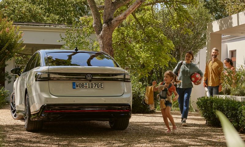 View of the rear of a VW ID.7 standing under trees in front of a house. A family with children runs towards it.