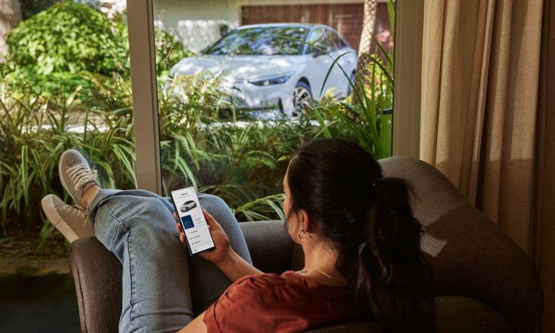 Una donna è seduta rilassata in salotto e guarda la Volkswagen ID.7 attraverso la finestra con uno smartphone in mano.