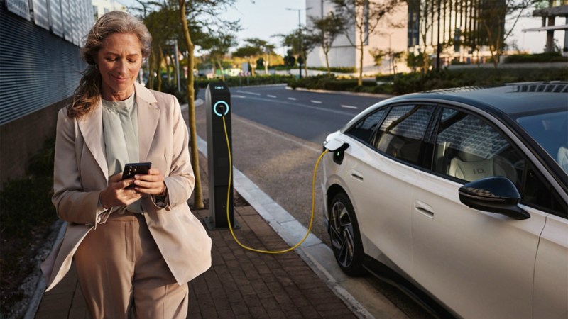 Side view of a white VW ID. charging at a public charging station. A woman passes in front of it, smartphone in hand.