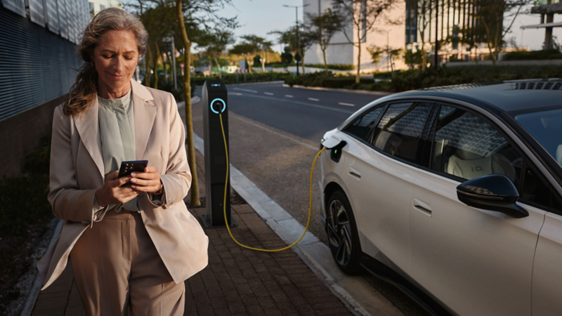 Eine Frau mit Smartphone geht neben einem weißen VW ID.7, der an einer Ladestation lädt.
