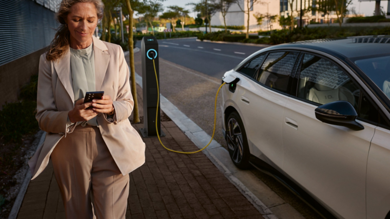 Eine Frau mit Smartphone geht neben einem weißen VW ID.7, der an einer Ladestation lädt.