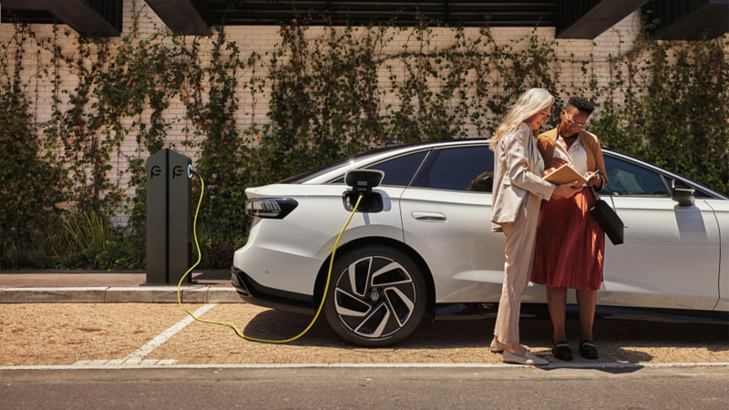 Two women standing with a book in hand next to a white VW ID. which is charging at a public charging station