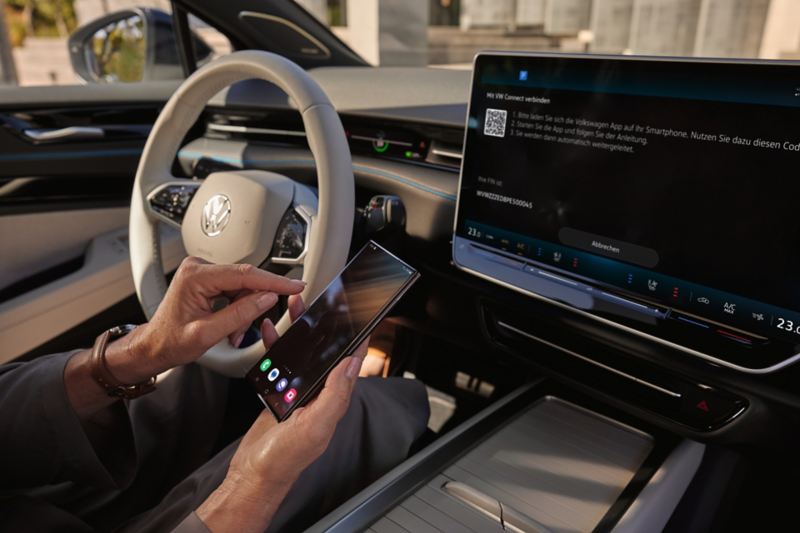 En person sitter i en Volkswagen og bruker Volkswagen-appen med en mobiltelefon