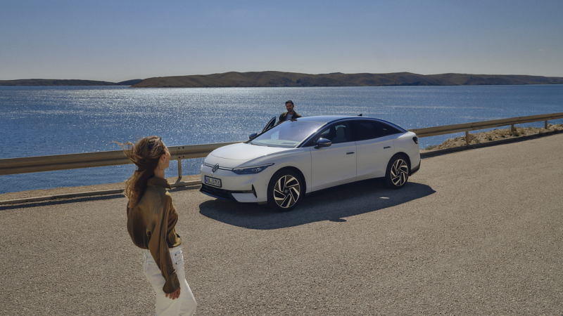 Biały Volkswagen ID.7 zaparkowany na drodze przy wybrzeżu. Mężczyzna otwiera drzwi samochodu. W tle woda, wzgórza i niebo. Na pierwszym planie idzie kobieta