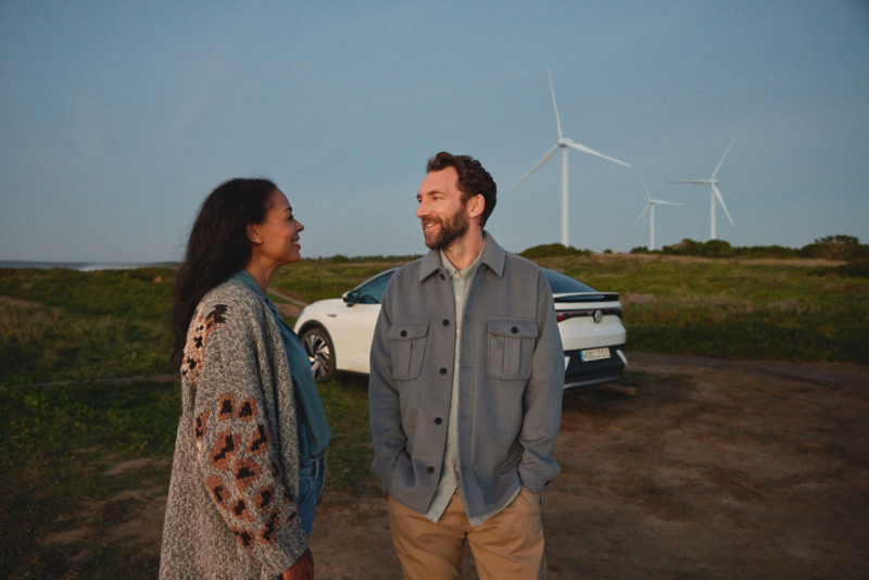 Un homme et une femme souriants se regardent, debout devant une Volkswagen ID.5 blanche garée, avec des éoliennes en arrière-plan sous un ciel dégagé.