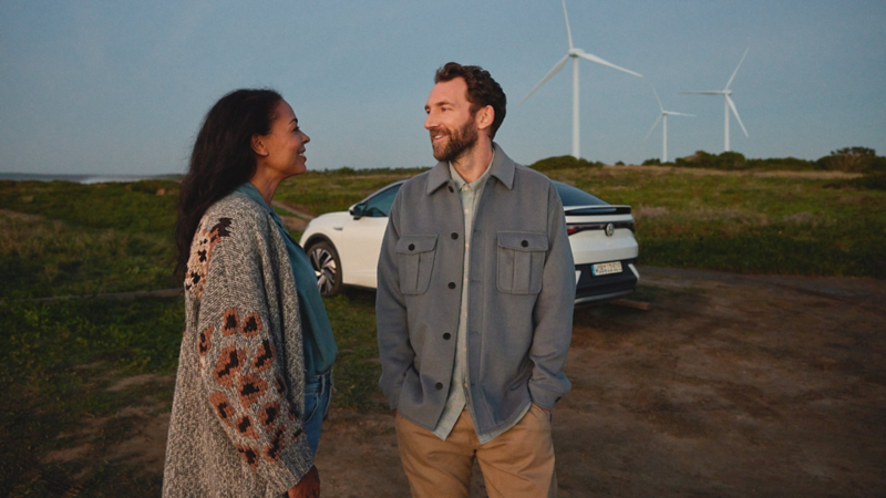 Eine Frau und ein Mann stehen vor einem weißen VW ID.5, der auf einem Feldweg parkt. Im Hintergrund Windräder.