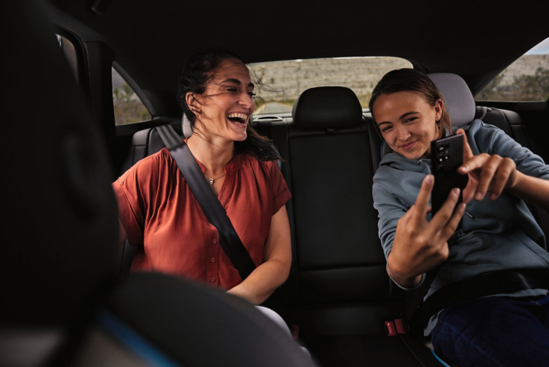  Deux passagers souriants sont assis sur la banquette arrière d'une voiture. La femme de gauche rit aux éclats, tandis que la jeune fille de droite prend un selfie ou une vidéo avec son téléphone.